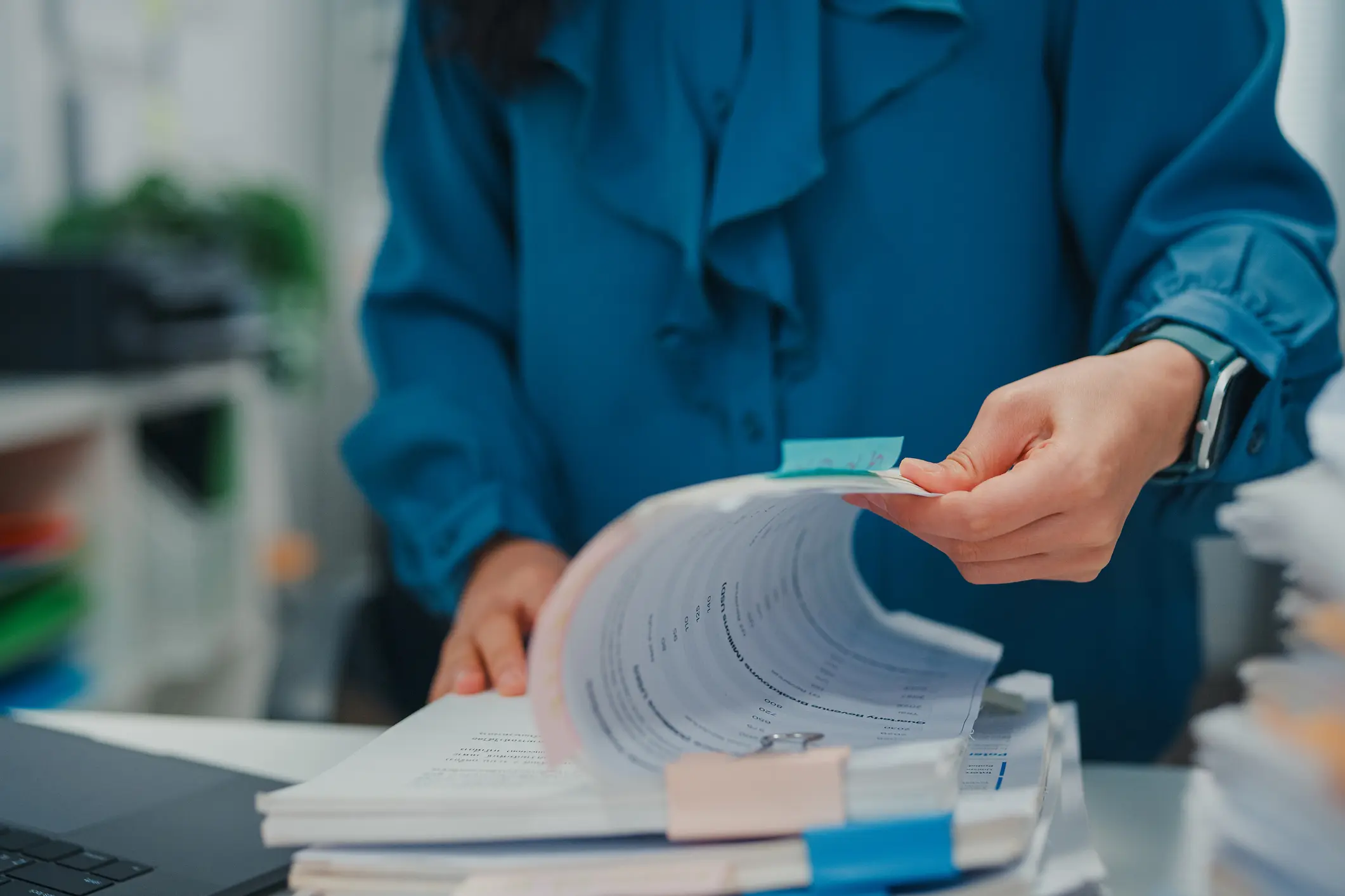 Closeup of overworked Asian businesswoman surrounded by piles of documents while working on laptop in busy office. Representing stress, workload, and modern corporate life concept.